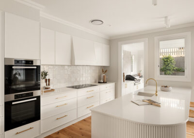 Modern white kitchen with a curved island, gold fixtures, built-in ovens, and an outdoor BBQ area
