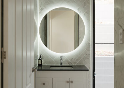 Custom bathroom vanity with white shaker cabinetry, stone benchtop, herringbone tiled wall and round backlit mirror