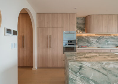 Coastal kitchen with arched bar nook, timber-look cabinetry, integrated ovens and stone splashback