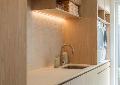 Laundry sink area with timber-look cabinetry, white benchtop and warm under-shelf lighting