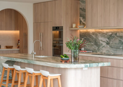Coastal kitchen with stone island, timber-look cabinetry, arched pantry way and feature splashback